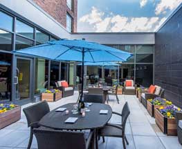 Image of Hyatt Place Hotel's enclosed open air patio with wooden flower boxes containing purple & yellow flowers, comfortable patio furniture with a bright blue umbrella and blue skies overhead, Harbor East Baltimore MD
